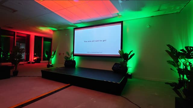 Large projection screen showing You are all set to go above a stage with red and green atmospheric lighting
